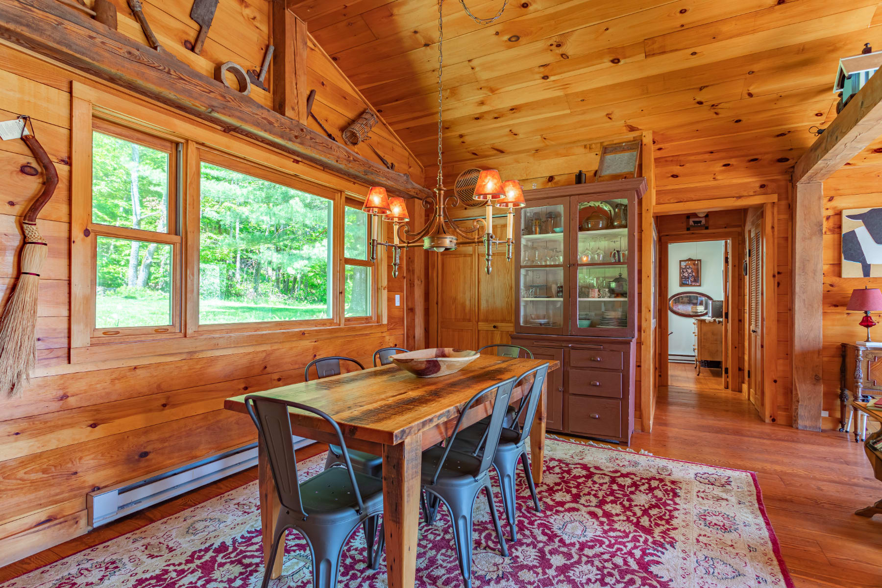 Rustic dining area with a long wooden farm table and black metal chairs, set on a patterned red rug, featuring exposed wooden walls and ceiling, and a vintage-style chandelier.