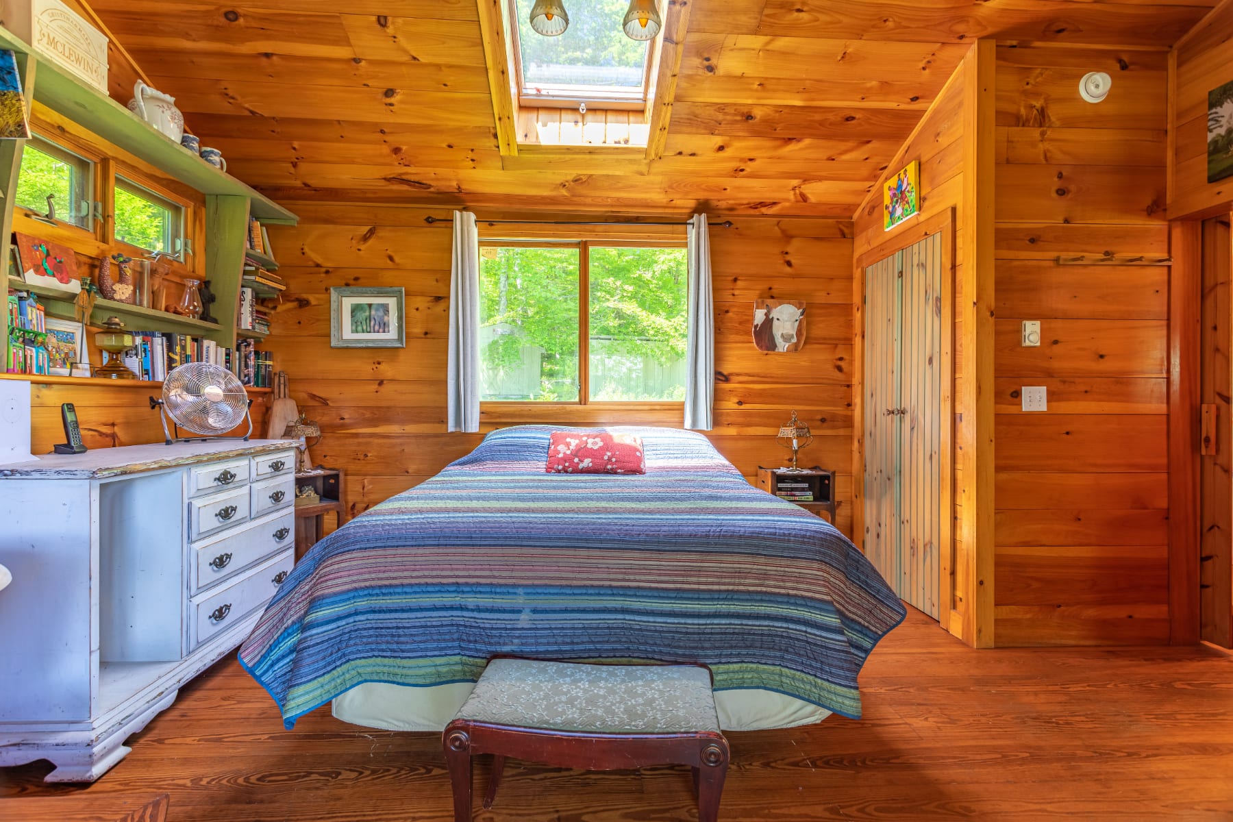 Warm wood-paneled bedroom featuring a striped bed with a colorful pillow, built-in shelving filled with books, and a skylight above, creating a bright and inviting space.