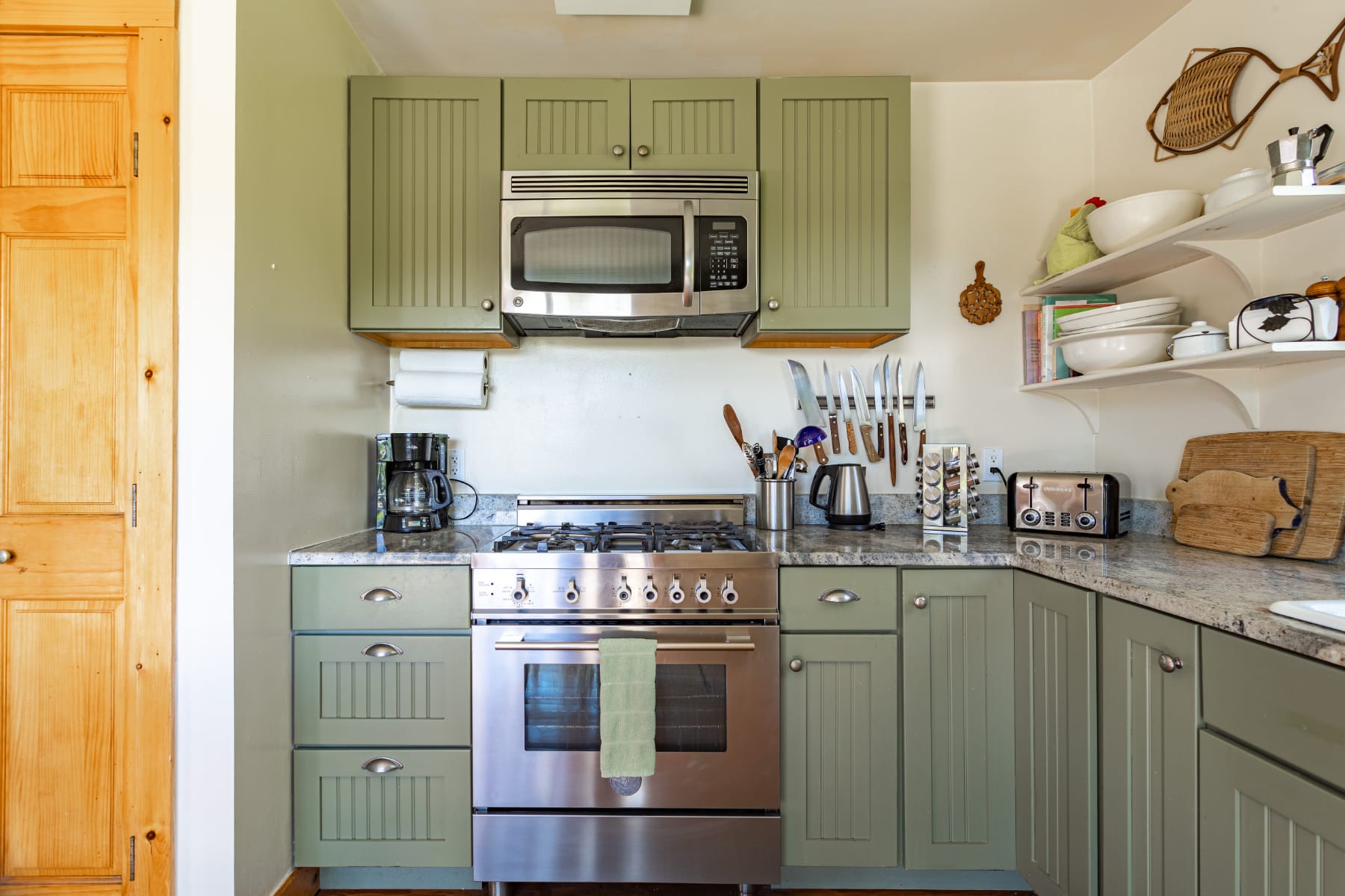Kitchen corner showcasing a stainless steel gas range with a microwave above, flanked by sage green shaker cabinets and light granite countertops, with a wooden pantry door to the left.