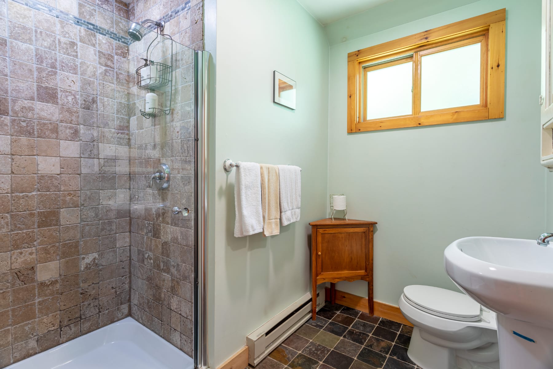 A light green bathroom featuring a tiled shower with a glass door, a white pedestal sink, and a toilet. A small wooden cabinet sits next to the toilet, and a window with a wooden frame provides natural light.