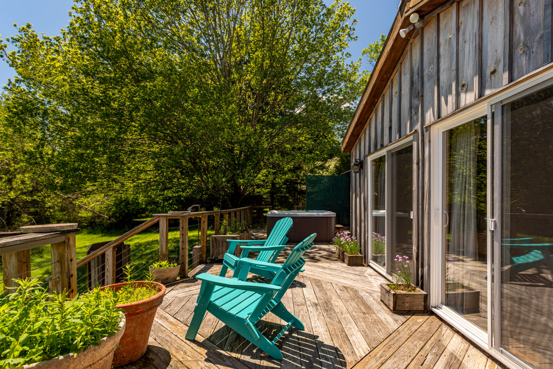 A rustic wooden deck extends from the cabin, featuring two turquoise Adirondack chairs and potted plants, with large sliding glass doors providing access to the interior and views of the surrounding greenery.