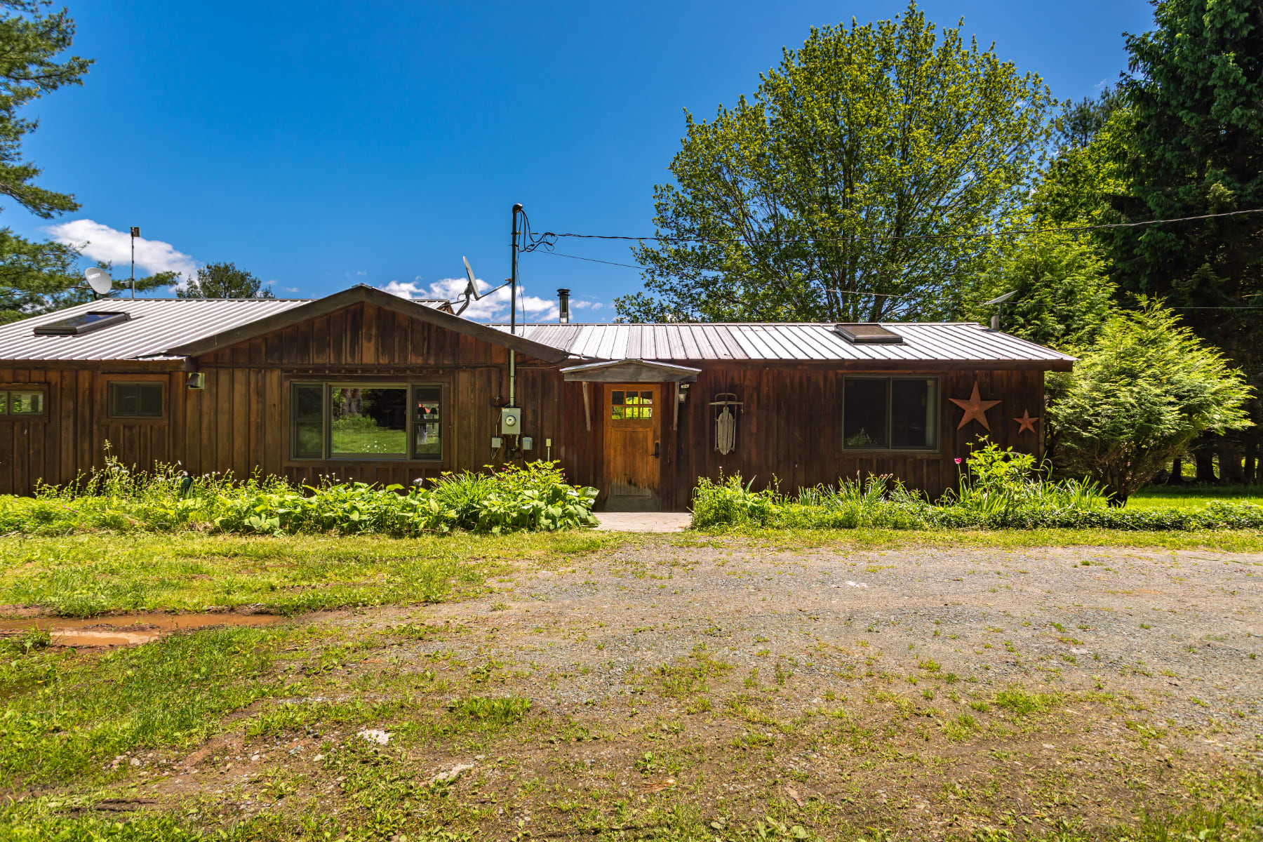 The front facade of a rustic wooden cabin with a metal roof, featuring a central wooden entry door, multiple windows, and a gravel driveway leading up to the entrance, surrounded by green foliage and trees.