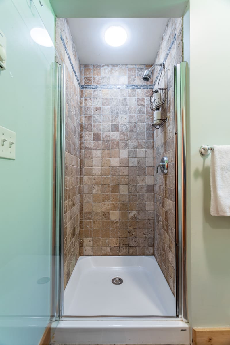 A close-up view of the shower in bathroom-1, featuring square brown and beige tiles, a glass enclosure, and a white shower pan. A circular skylight illuminates the shower stall from above.