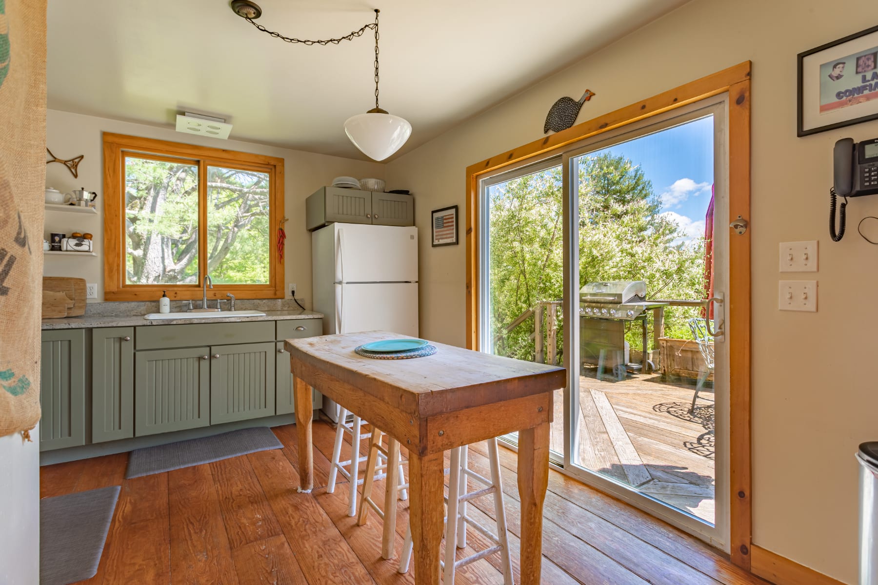 Bright kitchen featuring light gray cabinetry, a rustic wooden island with stools, and a white refrigerator, with a large sliding glass door opening to an outdoor deck.