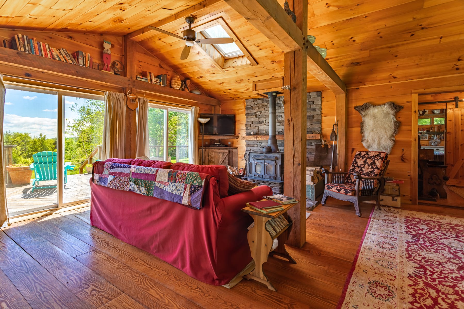 This bright living room boasts warm wooden walls and a vaulted ceiling, featuring a comfortable red sofa with a patchwork quilt, a wood-burning stove, and sliding glass doors that open to an outdoor deck with scenic views.