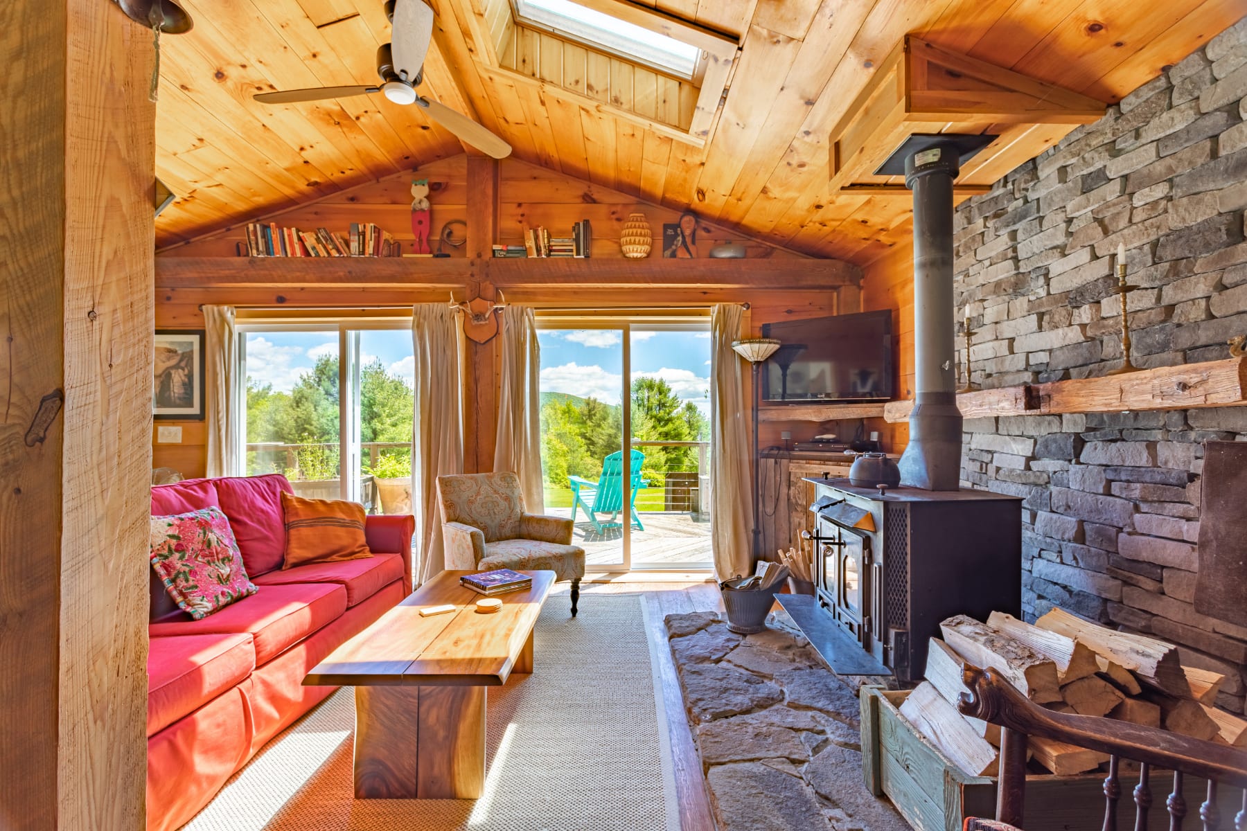 Bright living room with a stone fireplace and wood stove, featuring a vibrant red sofa and a wooden coffee table, with large sliding glass doors opening to a deck and offering scenic mountain views.