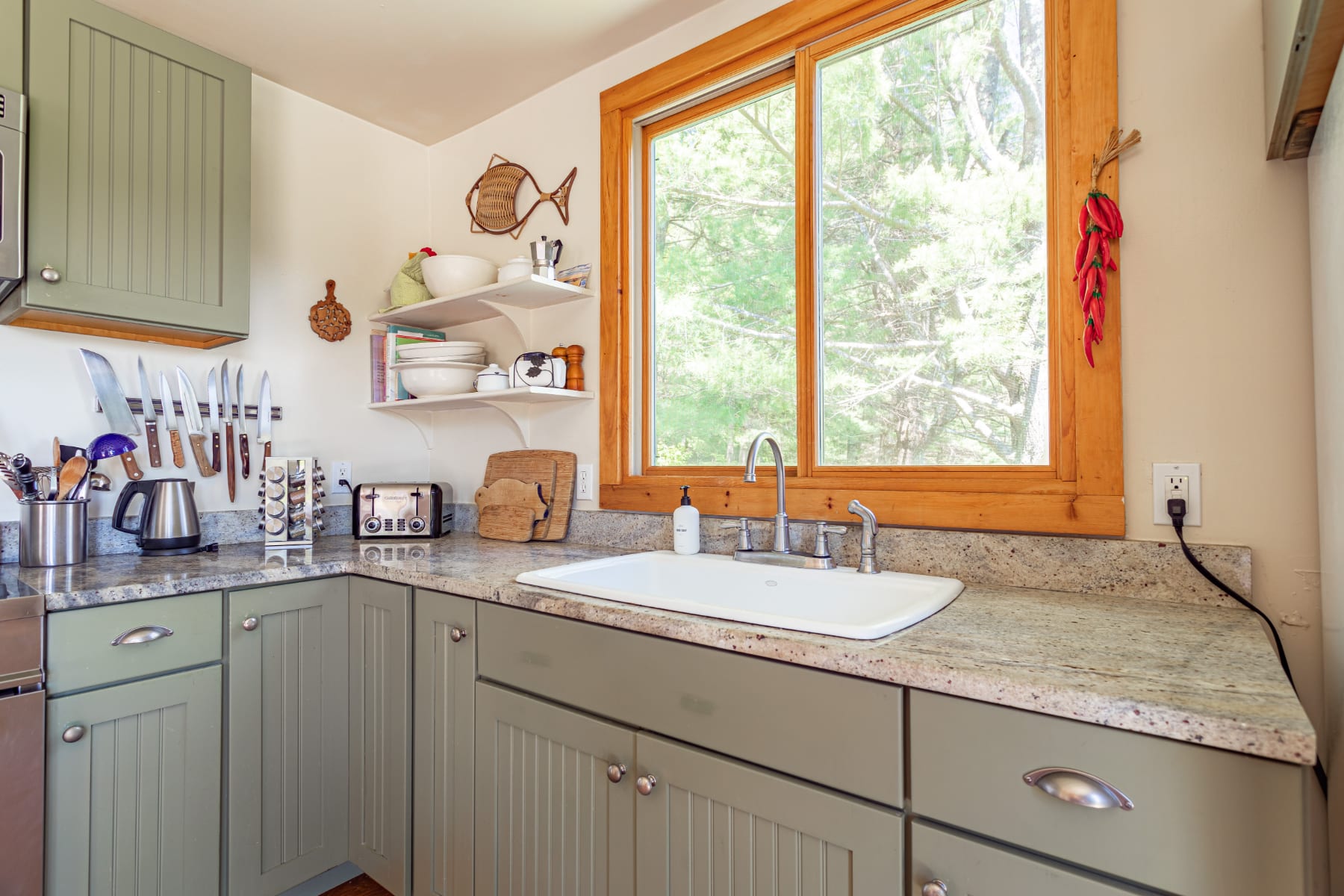 Close-up of the kitchen counter with a white farmhouse sink, light granite countertops, and open shelving displaying white dishes, illuminated by a large wooden-framed window.