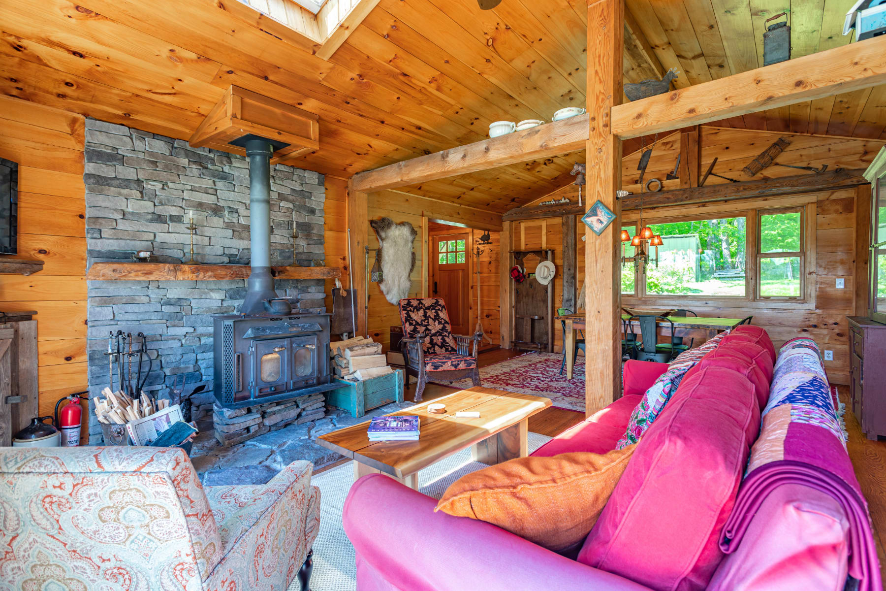 Cozy rustic living room with a prominent stone fireplace and wood-burning stove, exposed wooden beams, and a vaulted ceiling with a skylight, featuring a bright pink sofa and patterned armchair.