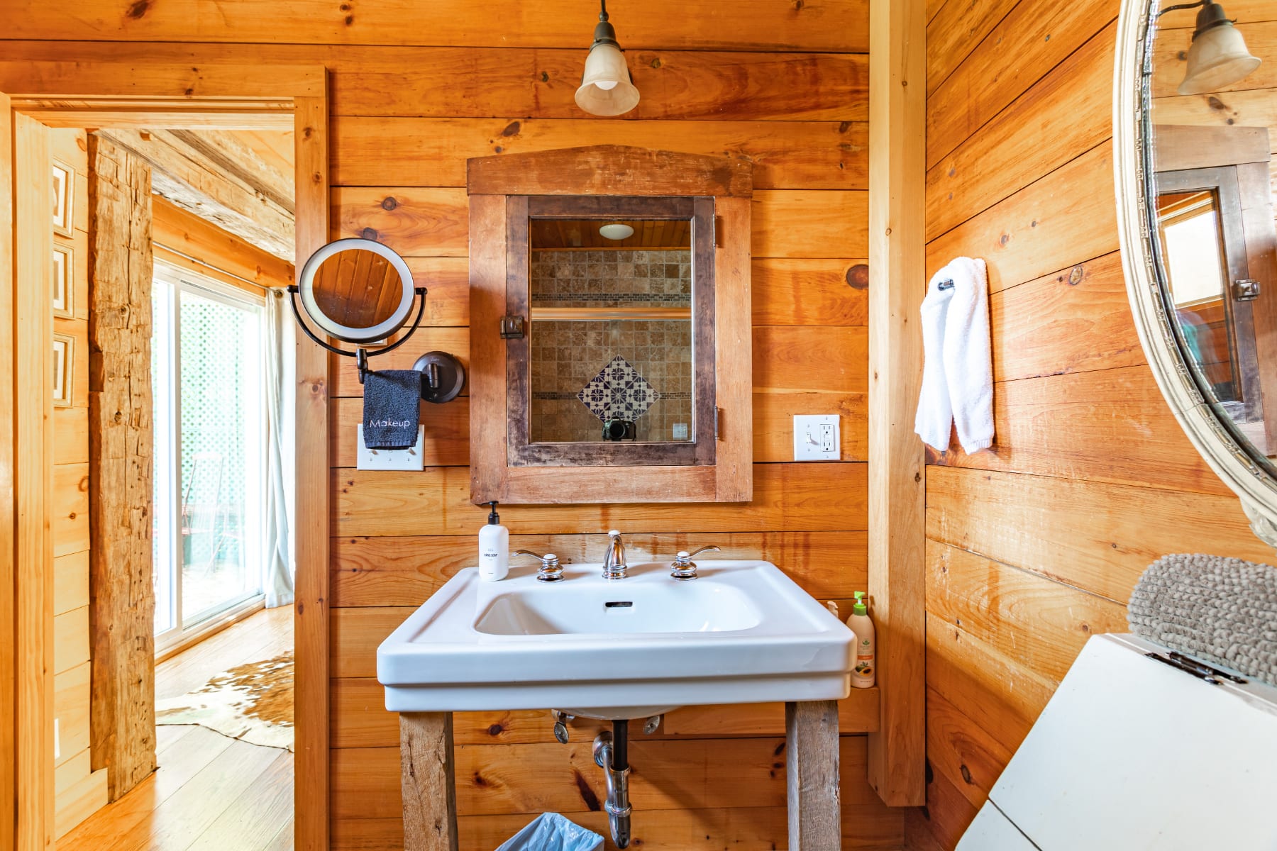 Bathroom vanity area with a white pedestal sink, a wooden medicine cabinet with a mirror, and an additional magnifying mirror, all set against warm wood-paneled walls.