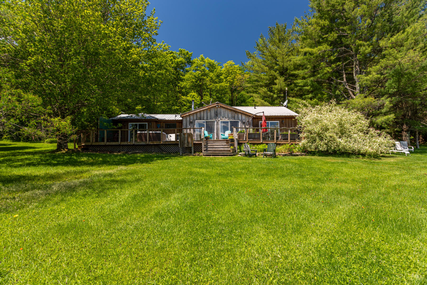 A rustic wooden cabin with a metal roof, featuring a large wraparound deck with outdoor seating and a hot tub, set against a backdrop of lush green trees and a vast grassy lawn under a clear blue sky.