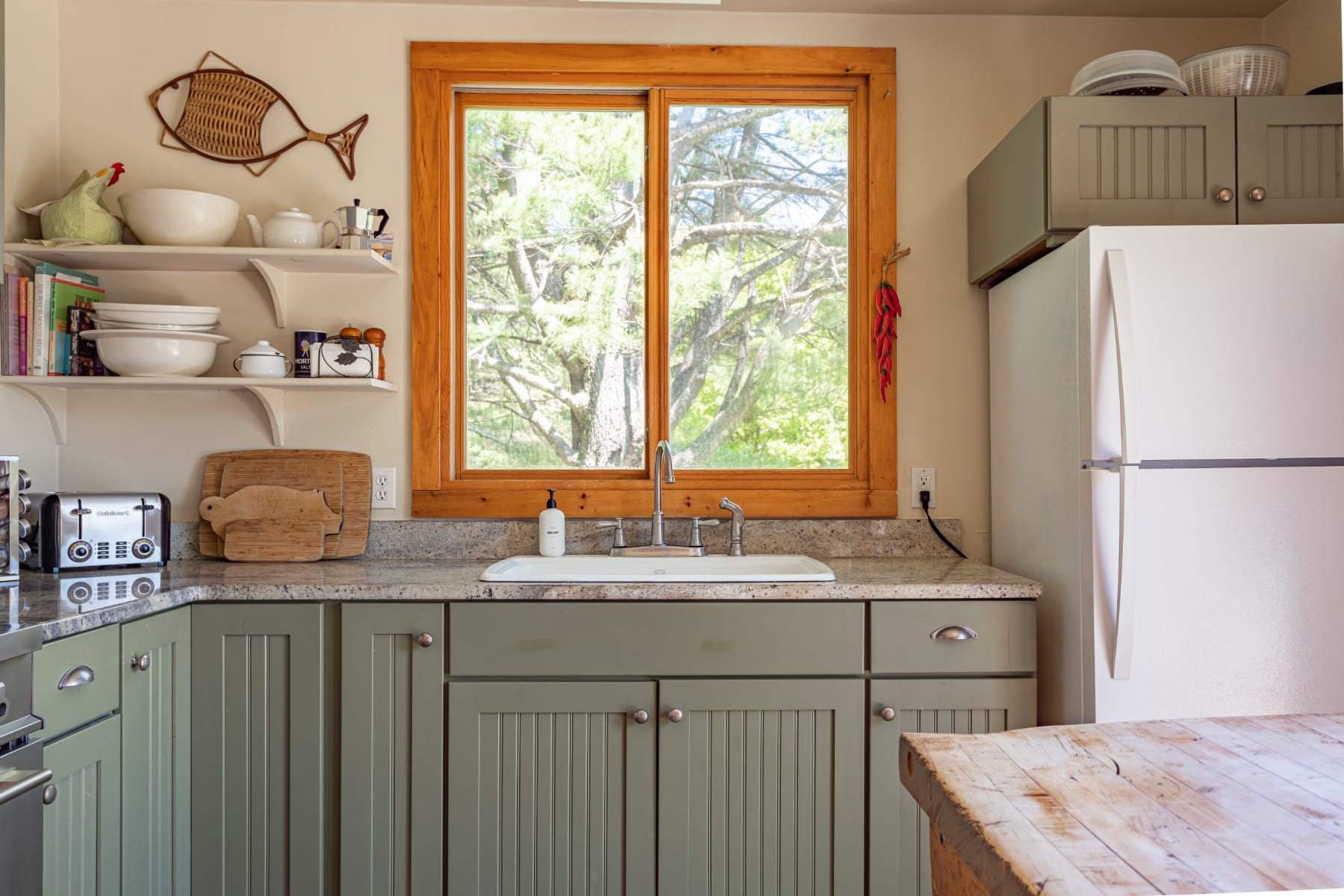 Detailed view of the kitchen sink area with light granite countertops, a white farmhouse sink, and a large wooden-framed window offering a view of lush trees, above sage green base cabinets.