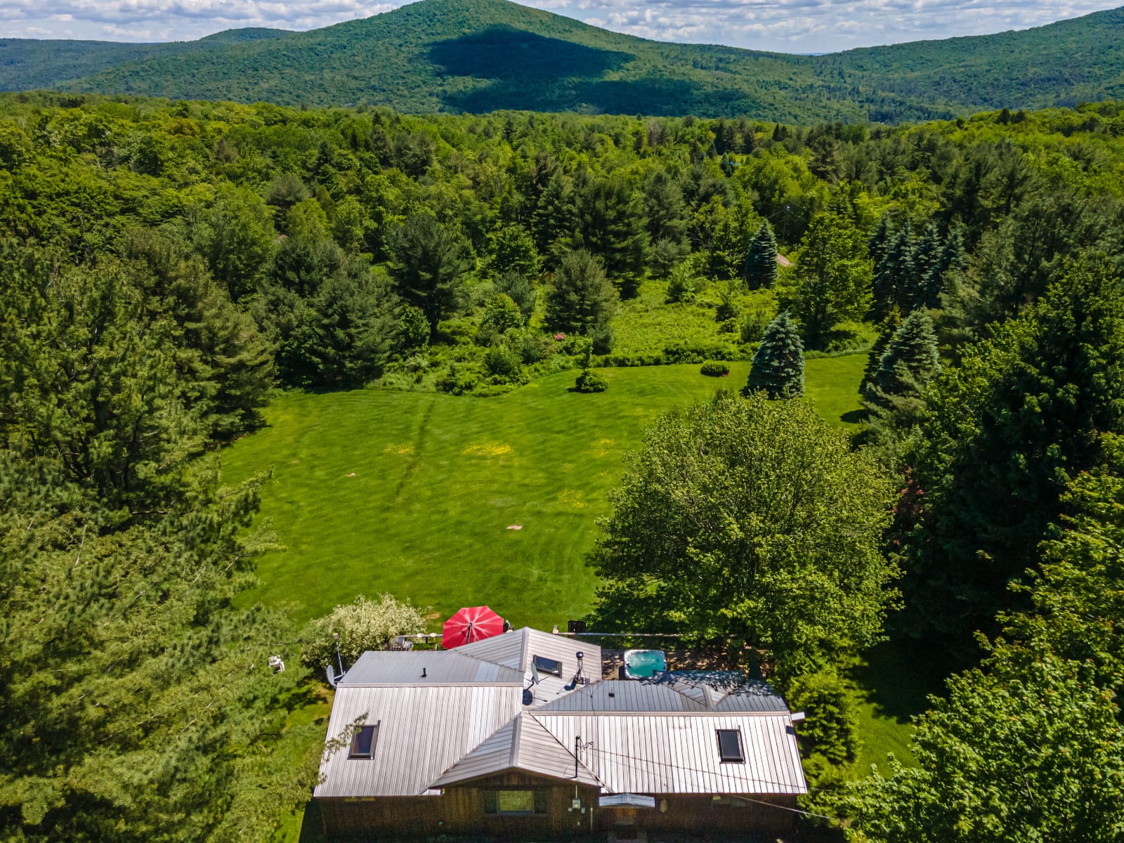 A high aerial view showcasing the rustic cabin nestled within a vast green property, surrounded by lush forests, with distant rolling mountains under a partly cloudy sky, highlighting the secluded natural setting.