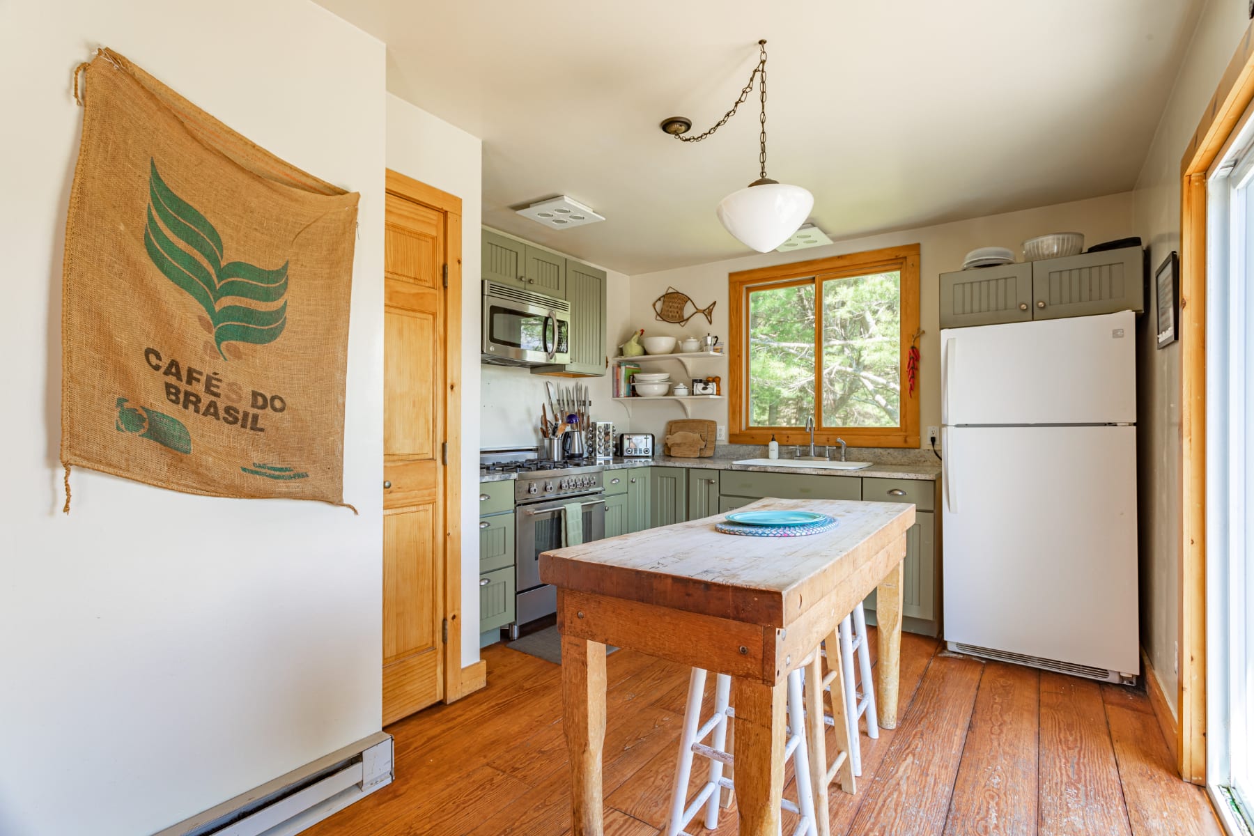 Bright, compact kitchen featuring sage green shaker-style cabinets, light granite countertops, and a wooden island with white stools, complemented by light wood floors and a large window overlooking greenery.