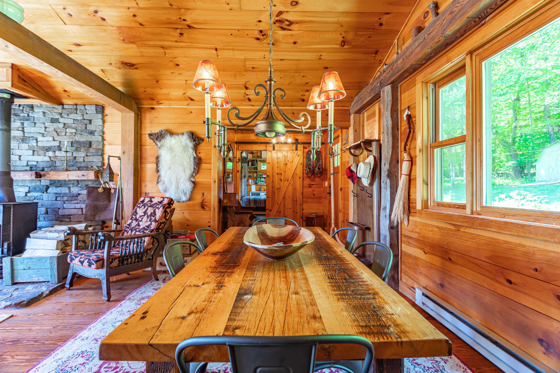 Spacious rustic dining room with a large wooden farm table, black industrial chairs, and a vintage chandelier, showcasing exposed wooden beams and walls, with a view towards the living area and a stone fireplace.