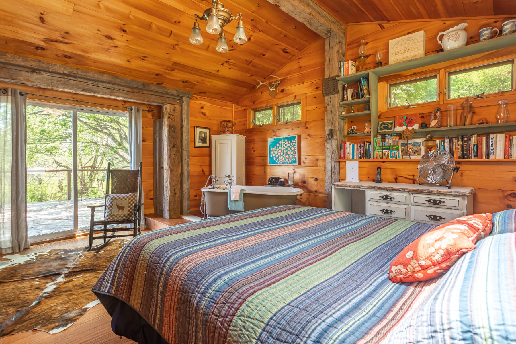 Cozy bedroom with warm wood-paneled walls and ceiling, featuring a striped bed, a freestanding clawfoot bathtub on a raised platform, and a sliding glass door leading to an outdoor deck.