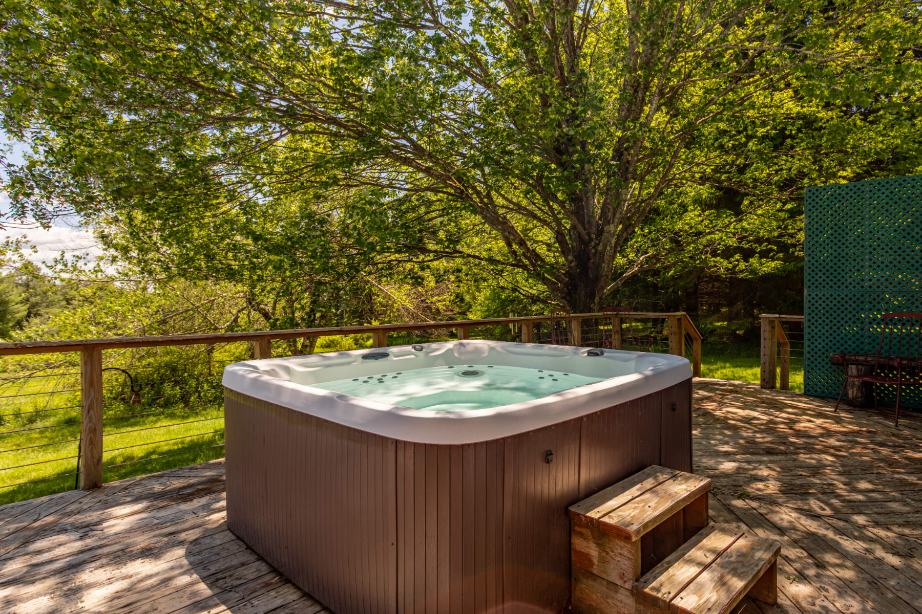 A large hot tub with wooden steps is positioned on a spacious wooden deck, surrounded by mature trees providing shade. The hot tub is filled with clear water, ready for use.