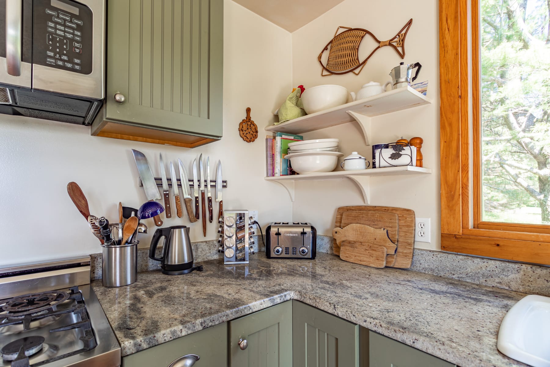 Kitchen counter detail showing a toaster, knife block, and kettle on light granite countertops, with sage green cabinets below and open shelving above, next to a large window.