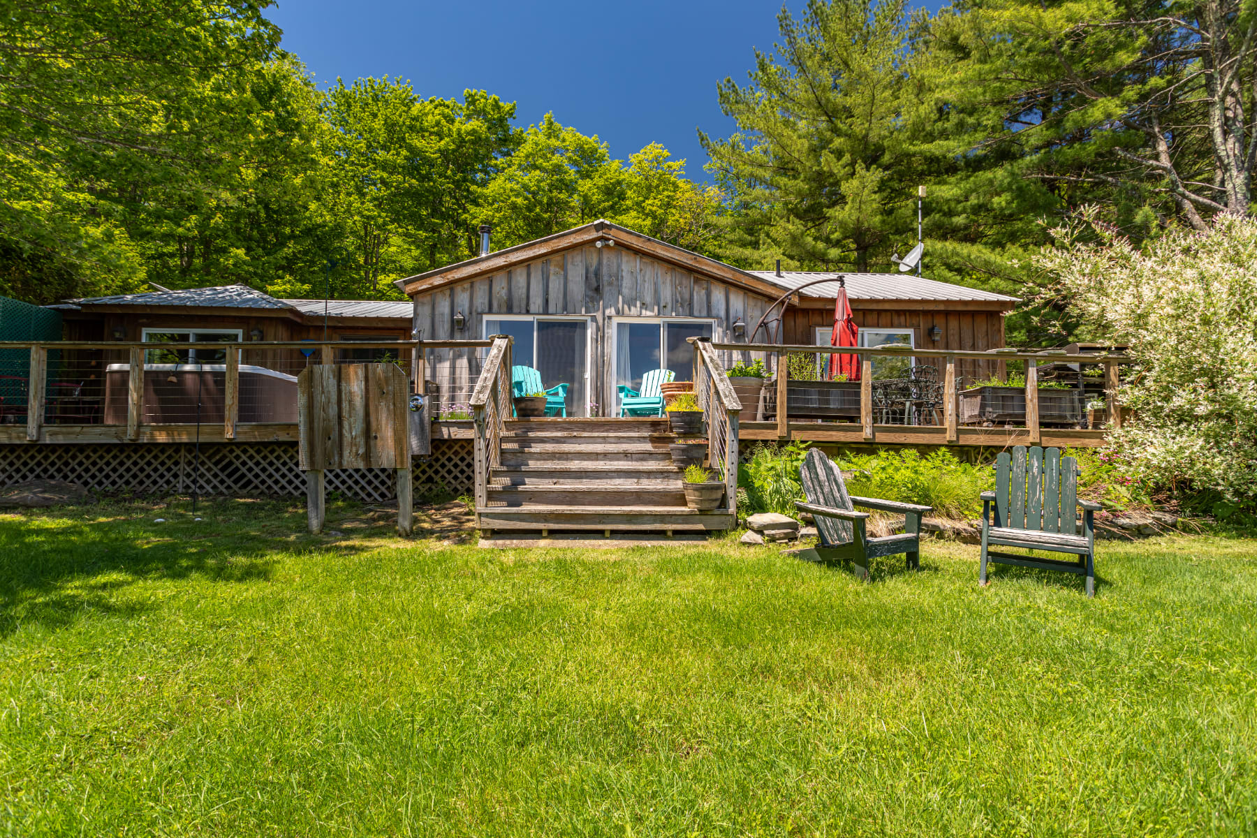 Rustic wooden cabin with a large wraparound deck featuring a hot tub, outdoor seating, and Adirondack chairs on a grassy lawn, surrounded by lush green trees under a clear blue sky.