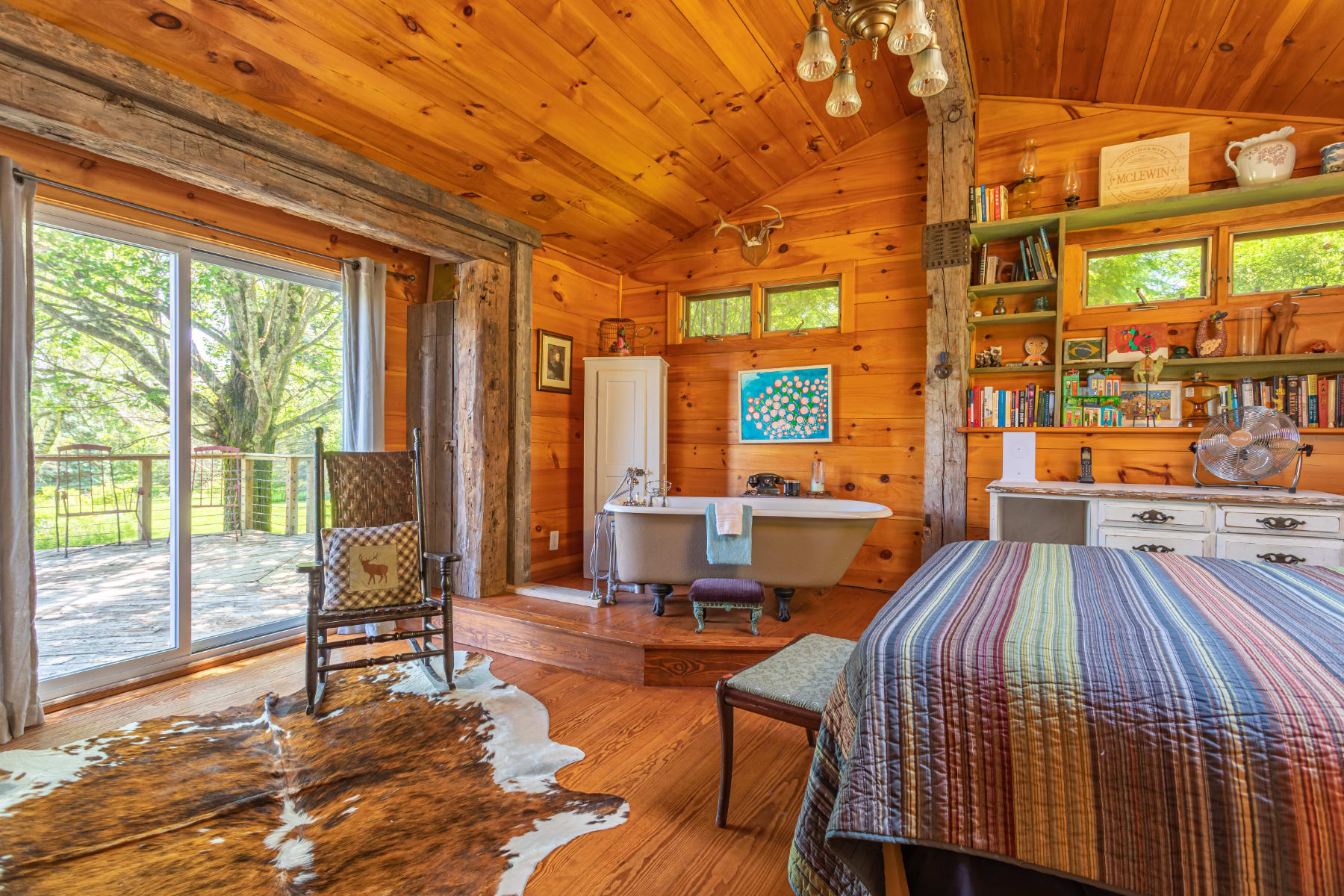 Rustic bedroom with extensive wood paneling, a comfortable striped bed, and a unique freestanding clawfoot bathtub positioned on a raised wooden platform, with a rocking chair nearby.