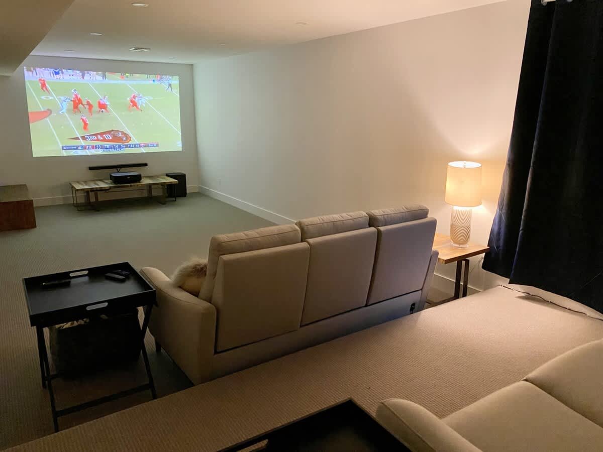 A cozy media room features a large projected screen displaying a football game, comfortable light-colored sofas, and a side table with a lamp, creating an inviting entertainment space.