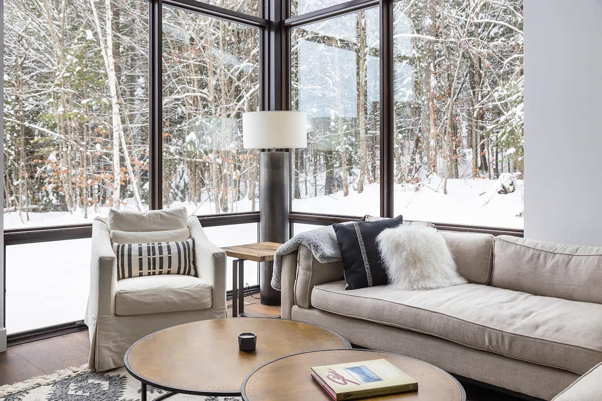 A bright living room features a comfortable sofa and armchair, a tall floor lamp, and expansive windows offering a serene view of a snow-covered forest, creating a peaceful ambiance.