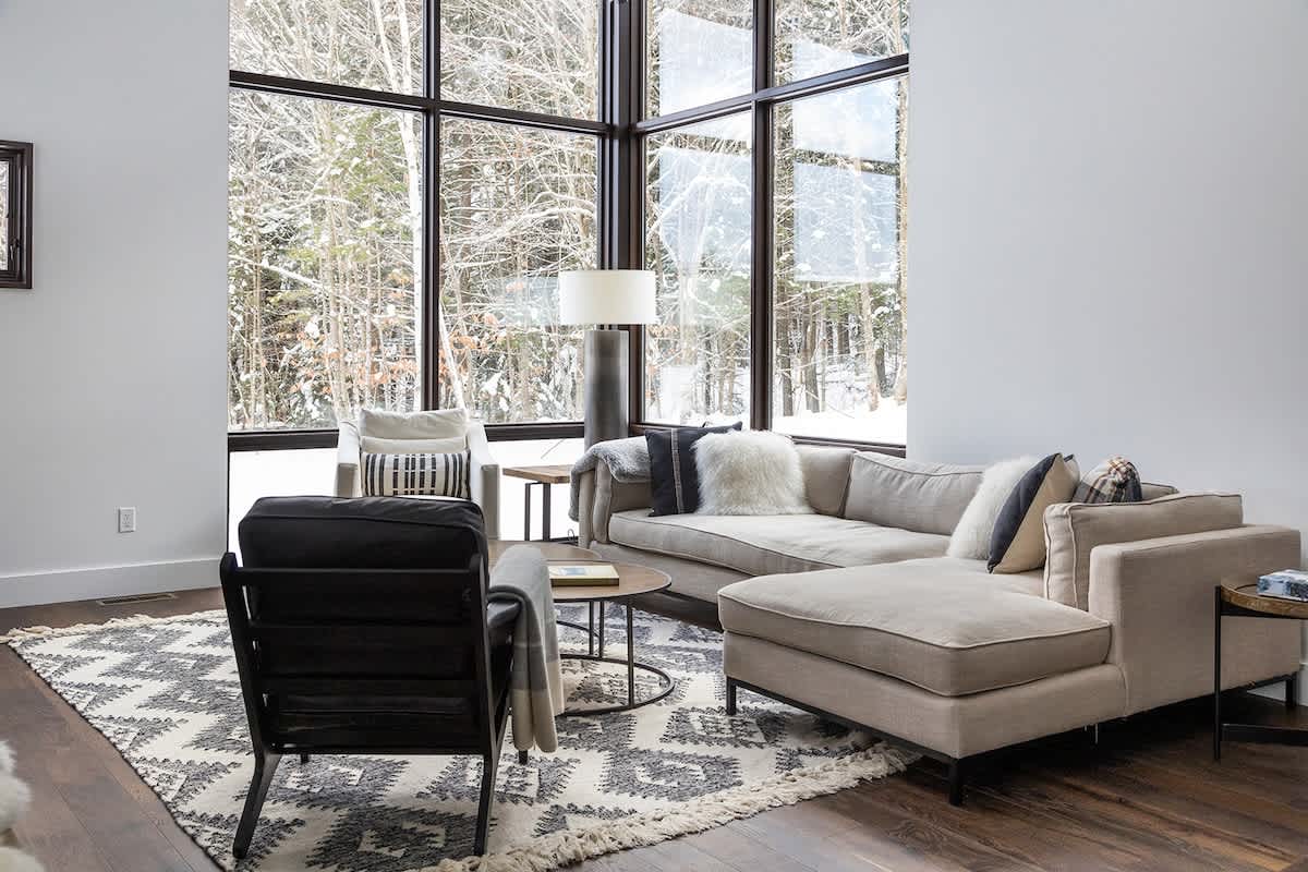 A spacious living room with a light-colored sectional sofa, a black armchair, and large floor-to-ceiling windows showcasing a snowy forest view, anchored by a patterned rug.