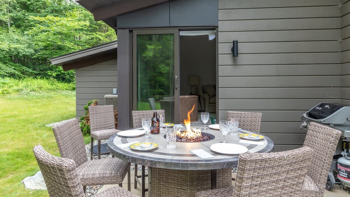 An outdoor patio area with a round dining table featuring a built-in fire pit, surrounded by wicker chairs, with a BBQ grill visible and a green lawn leading to a forest.