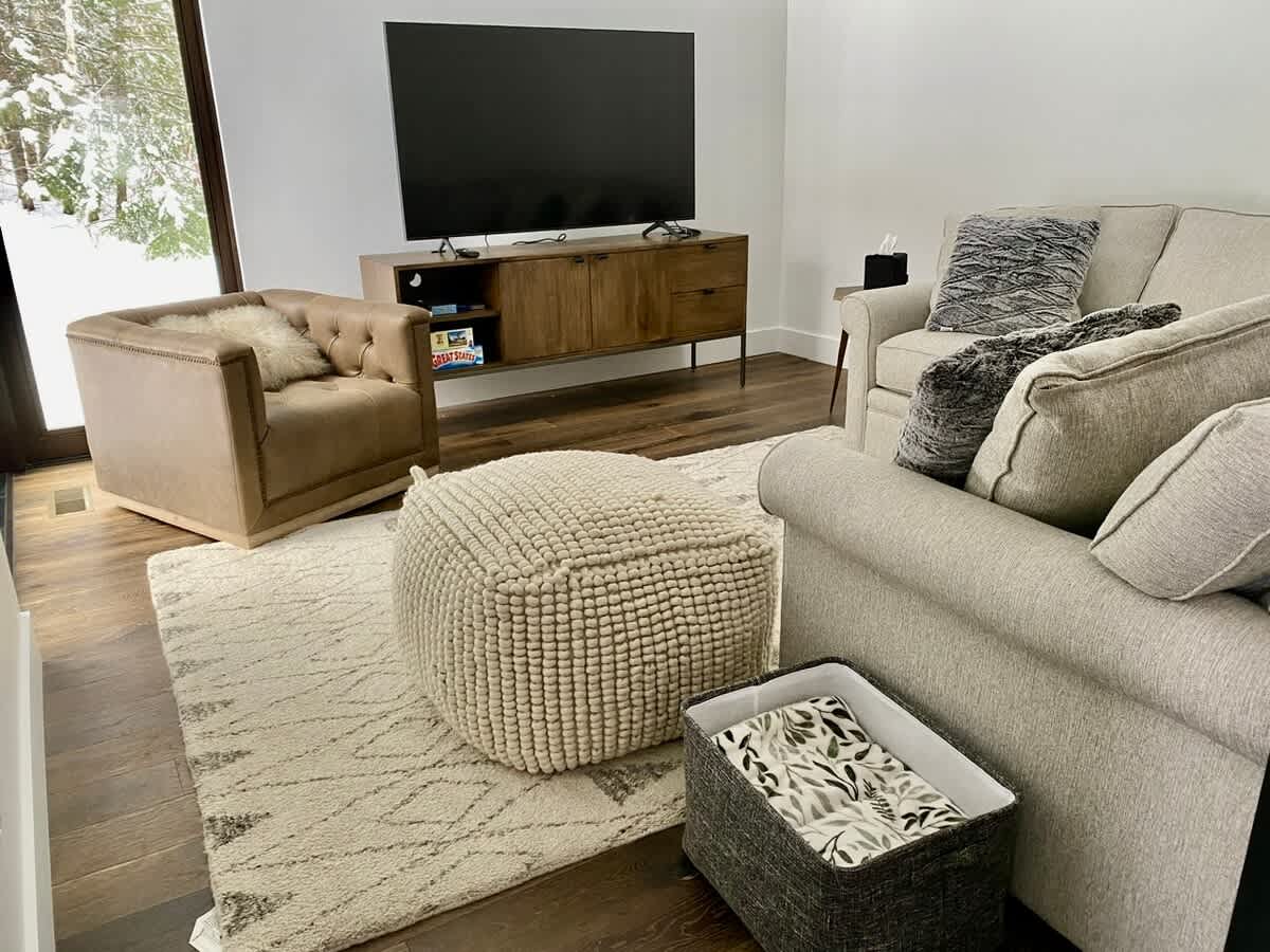 Cozy living room with a light-colored sofa, a beige tufted armchair, a large flat-screen TV on a wooden console, and a textured cream-colored pouf on a patterned rug, with dark hardwood floors.
