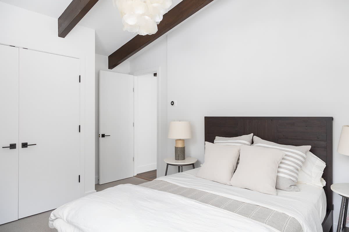 Modern bedroom with a dark wooden headboard, white bedding, and light gray walls. Exposed dark wooden beams accent the white ceiling, and a bedside lamp sits on a small round table.