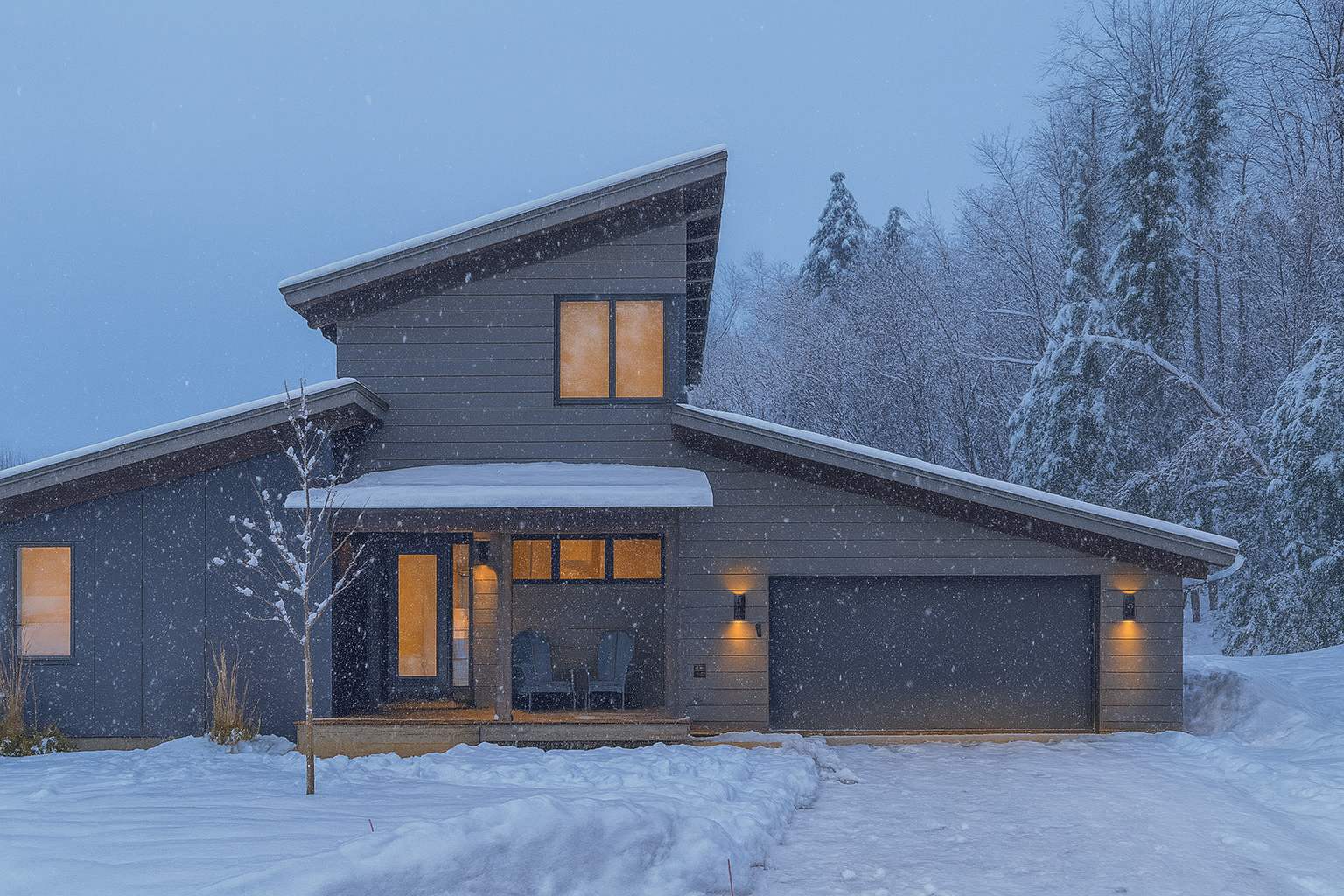 A modern house with a distinctive sloped roofline and gray siding, illuminated at dusk during a snowfall, with a snow-covered driveway and surrounding trees.