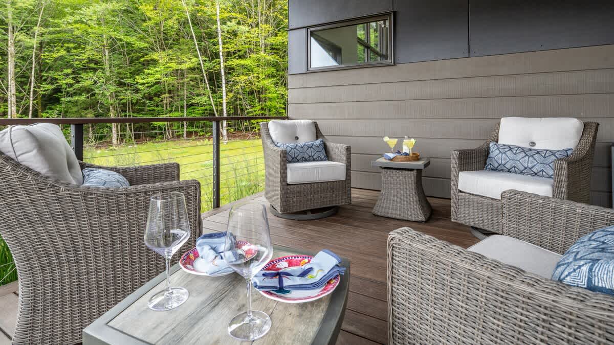 A spacious outdoor deck with several wicker armchairs and a small table, overlooking a lush green forest, with wine glasses and plates set on a coffee table.