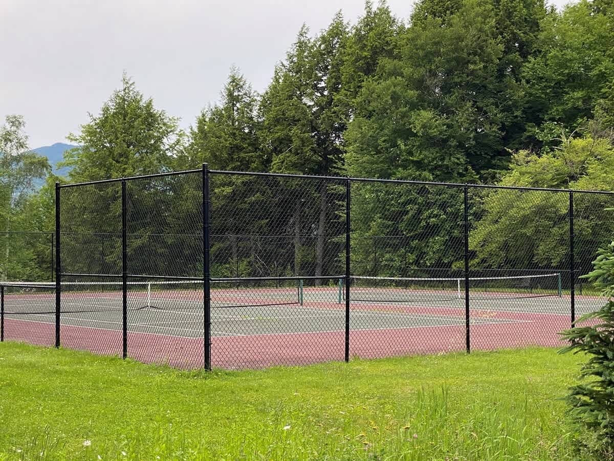 Outdoor tennis court with a green and red surface, enclosed by a black chain-link fence, surrounded by dense green trees and distant mountains under a cloudy sky.