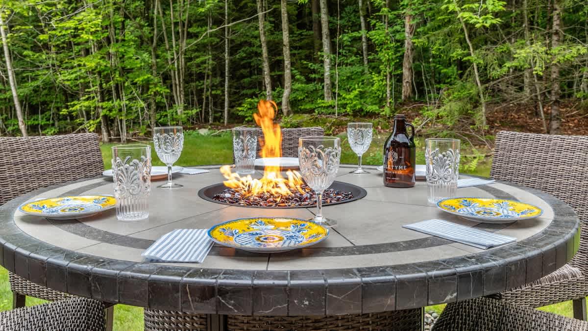 An inviting outdoor dining area features a round table with a built-in fire pit, set with plates and glasses, surrounded by wicker chairs and lush green trees in the background.