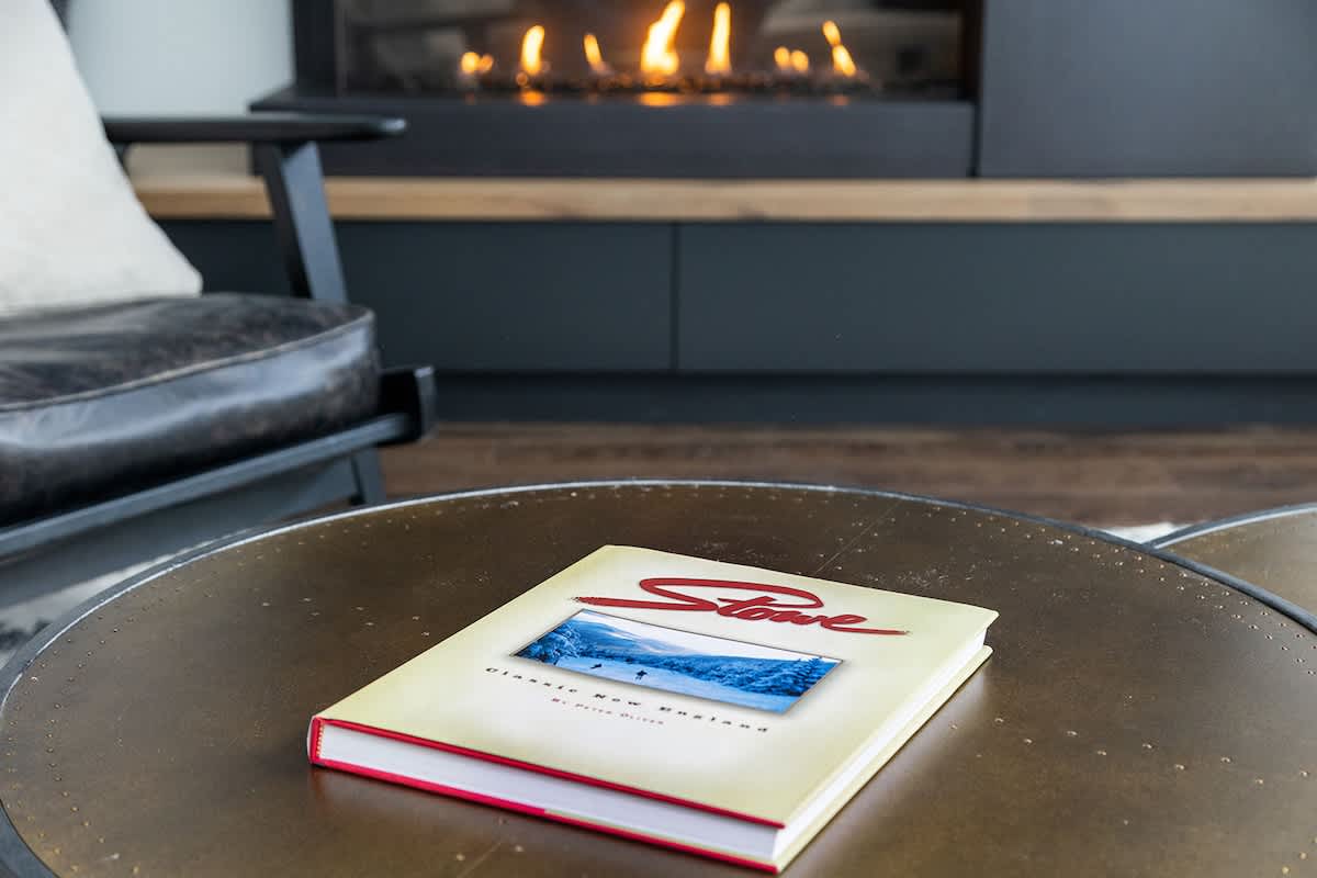 A close-up of a round metal coffee table with a book titled "Stowe" on it, positioned in front of a modern fireplace with visible flames, adding a cozy touch to the living space.