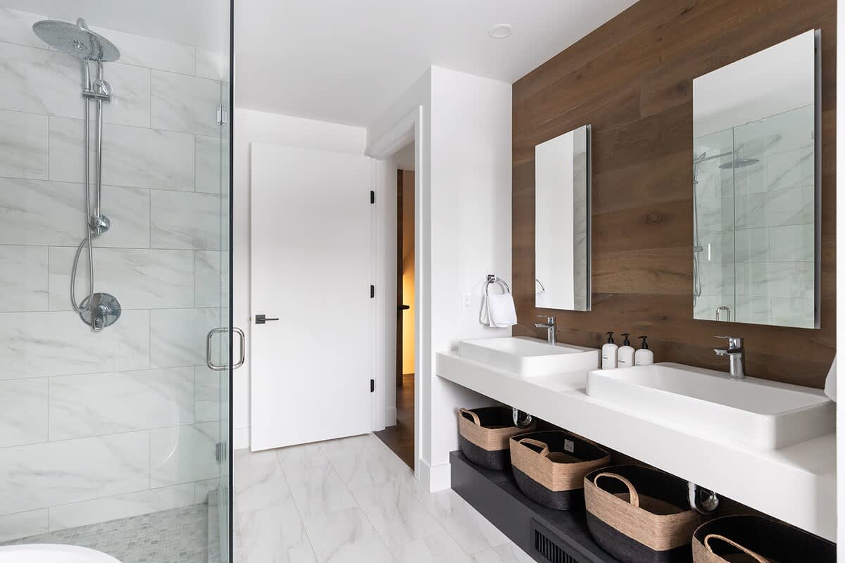 Modern bathroom featuring a glass-enclosed shower with white tiled walls, a double vanity with white rectangular sinks, and a wooden accent wall behind the mirrors, with light-colored floor tiles.