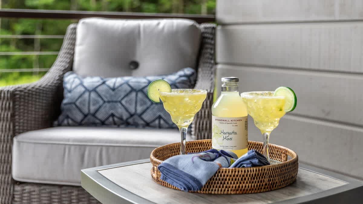 A close-up of a wicker outdoor chair with a patterned pillow, and a small table holding a tray with two margarita glasses and a bottle of margarita mix.