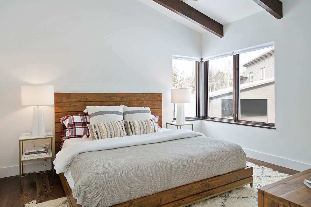 Bright bedroom with a wooden headboard and bed frame, white bedding with plaid accents, two white table lamps on gold nightstands, and large corner windows overlooking a snowy outdoor scene.