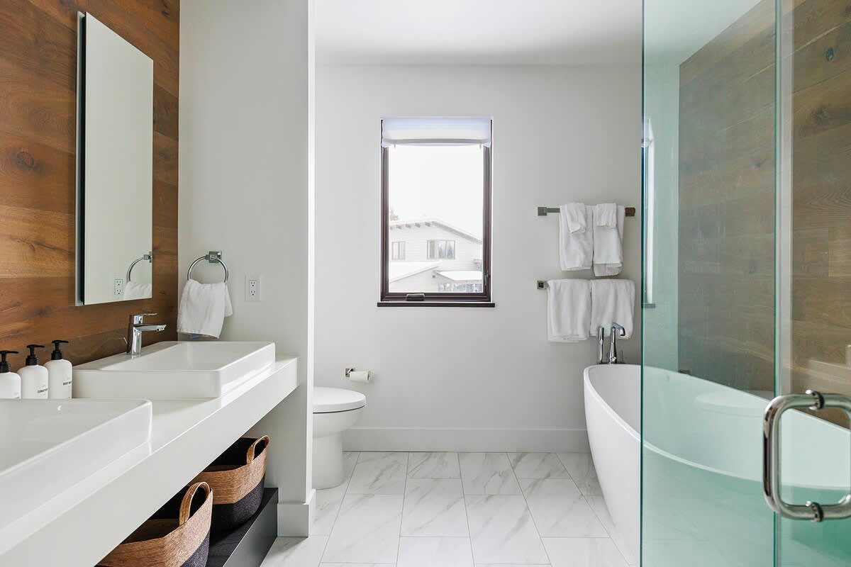 Contemporary bathroom with a freestanding bathtub, a toilet, a double vanity with white sinks, and a wooden accent wall, featuring a large window and white tiled floors.