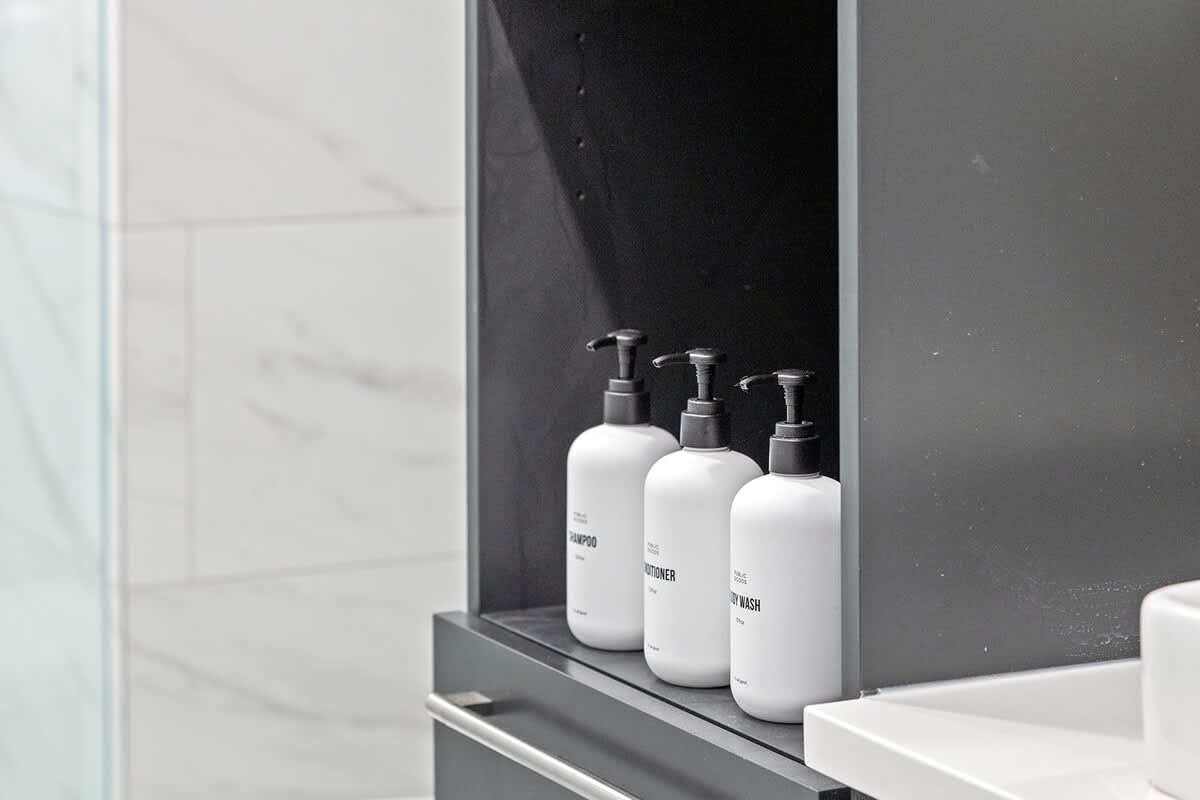 Close-up of a dark gray bathroom storage cabinet displaying three white pump bottles labeled "shampoo," "conditioner," and "body wash," alongside neatly folded white towels.