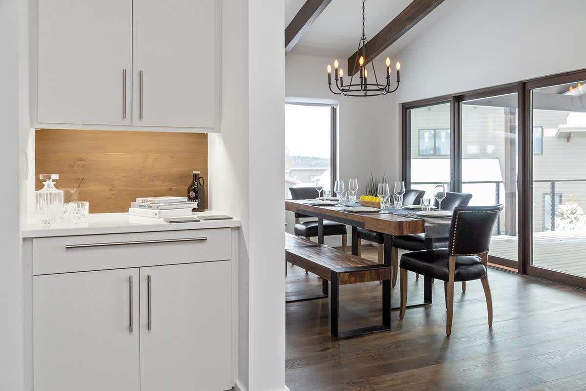 Dining area with a rustic wooden table, black leather chairs, a wooden bench, and a black chandelier, adjacent to large sliding glass doors and a built-in white cabinet with a wooden backsplash.