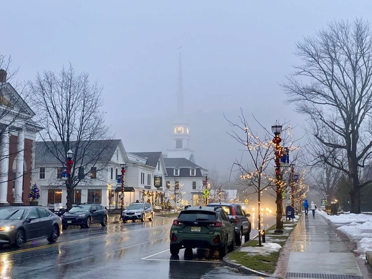 A foggy street scene in a small town, featuring white colonial-style buildings, parked cars, bare trees with string lights, and a prominent church steeple in the misty background.