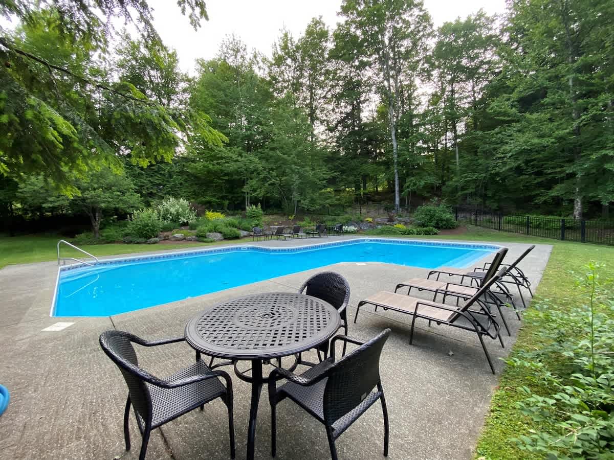 A large rectangular swimming pool surrounded by a concrete patio with lounge chairs and a round dining table with chairs, set amidst a lush green backyard with mature trees.