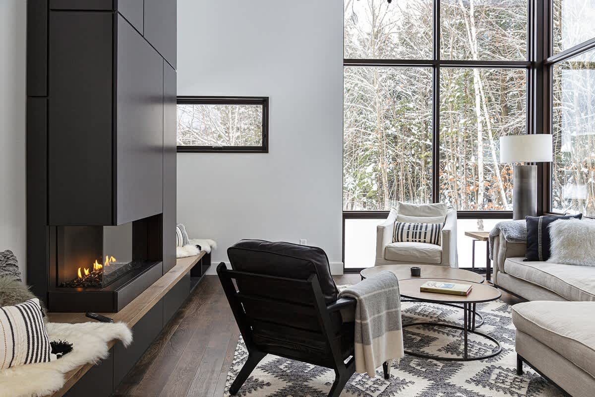A modern living room features a tall, dark fireplace with a built-in bench, a black leather armchair, and large windows offering serene views of a snowy landscape.