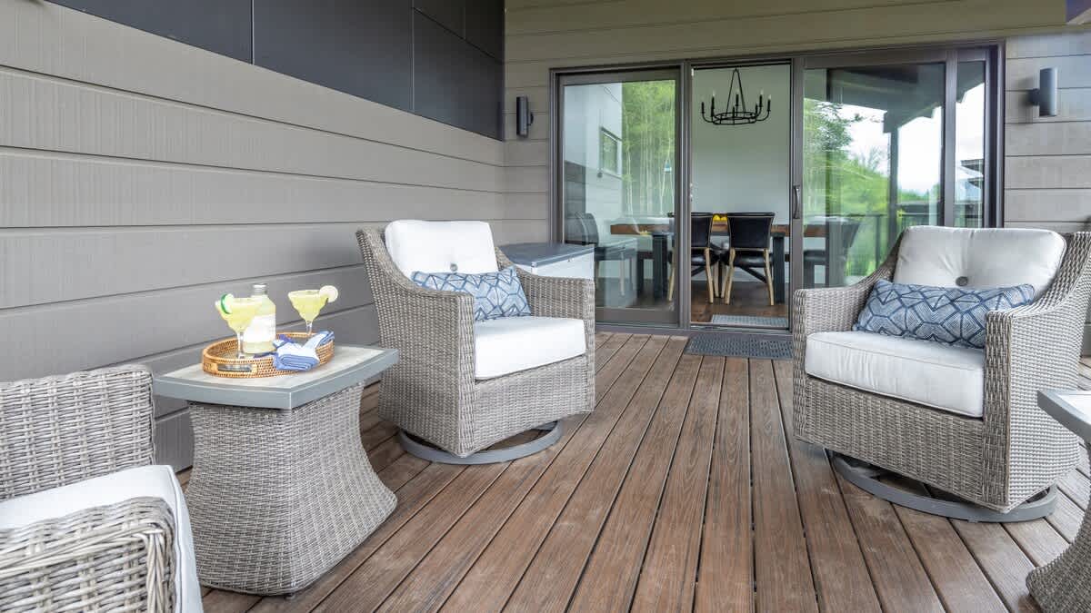 A modern outdoor deck with gray wicker swivel chairs and a side table, positioned in front of large sliding glass doors that lead into the house, with a view of the interior dining area.