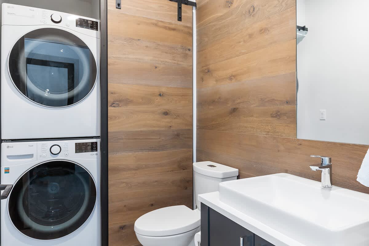 A modern bathroom featuring a stacked white washer and dryer unit next to a toilet, with a white vessel sink on a dark vanity, and a warm wood-paneled accent wall.