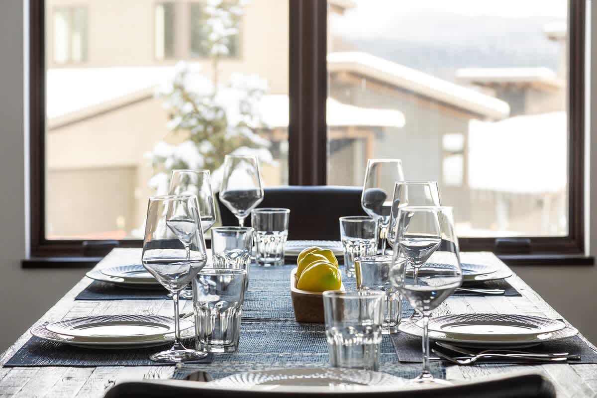 Close-up of a dining table set with plates, glasses, and cutlery, featuring a blue patterned runner and a bowl of lemons, with a snowy outdoor view through a large window in the background.