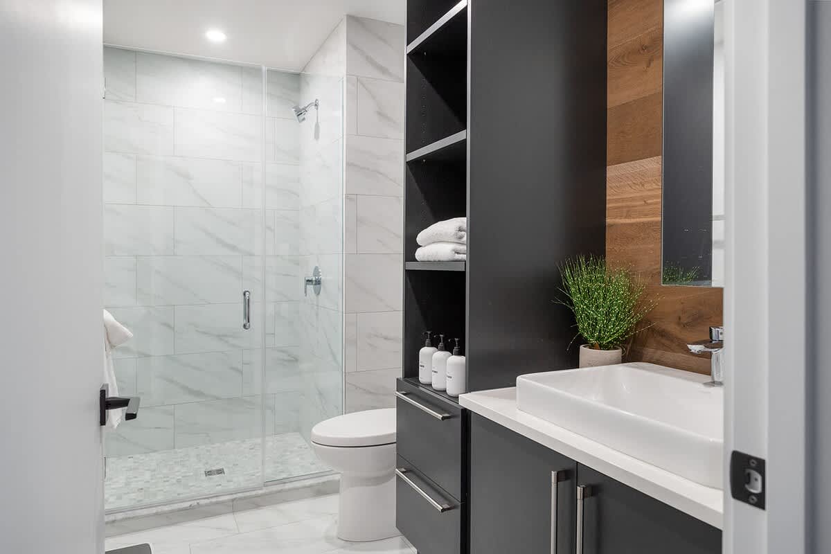 Contemporary bathroom with a white marble-tiled shower, a dark gray vanity with a white vessel sink, and a tall storage cabinet. A warm wooden accent wall adds contrast.