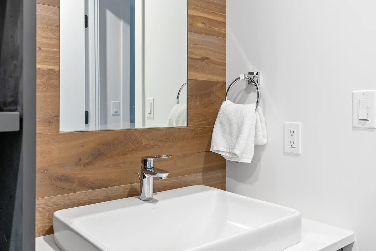 Detail of a modern bathroom vanity featuring a white vessel sink, chrome faucet, and a mirror set against a warm wooden accent wall. A white hand towel hangs nearby.