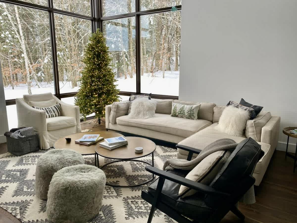 Cozy living room with a large sectional sofa, armchairs, and round coffee tables on a patterned rug, featuring a lit Christmas tree by expansive windows overlooking a snowy forest.