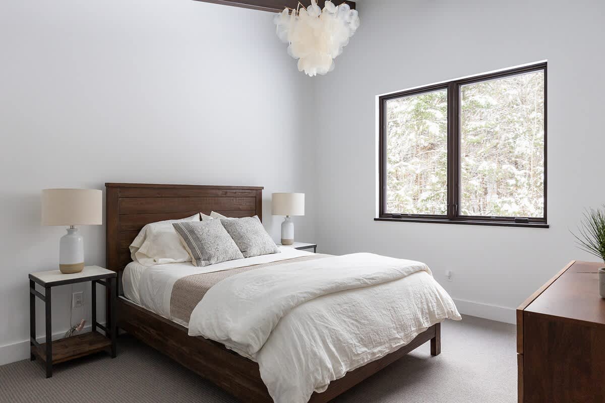 Bright bedroom with a wooden bed frame and headboard, flanked by two bedside tables with lamps. A large window offers a view of snowy trees, complementing the white walls and light gray carpet.